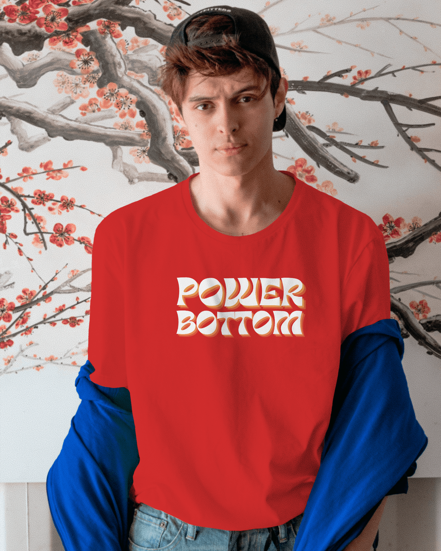 Person wearing a red sweatshirt with 'POWER BOTTOM' text against a floral wall. Kink, LGBTQIA+, Queer Sub Dom
