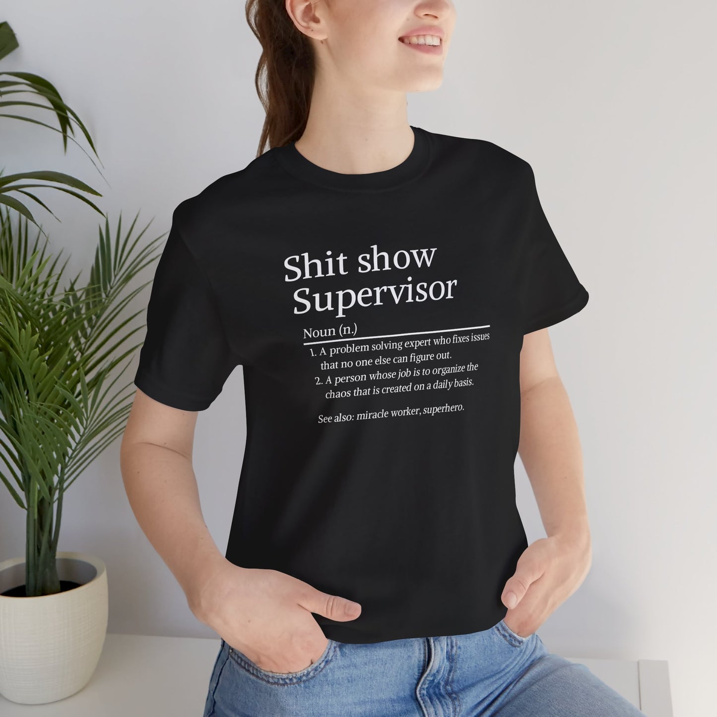 Person wearing a black t-shirt with text, standing indoors next to a plant.