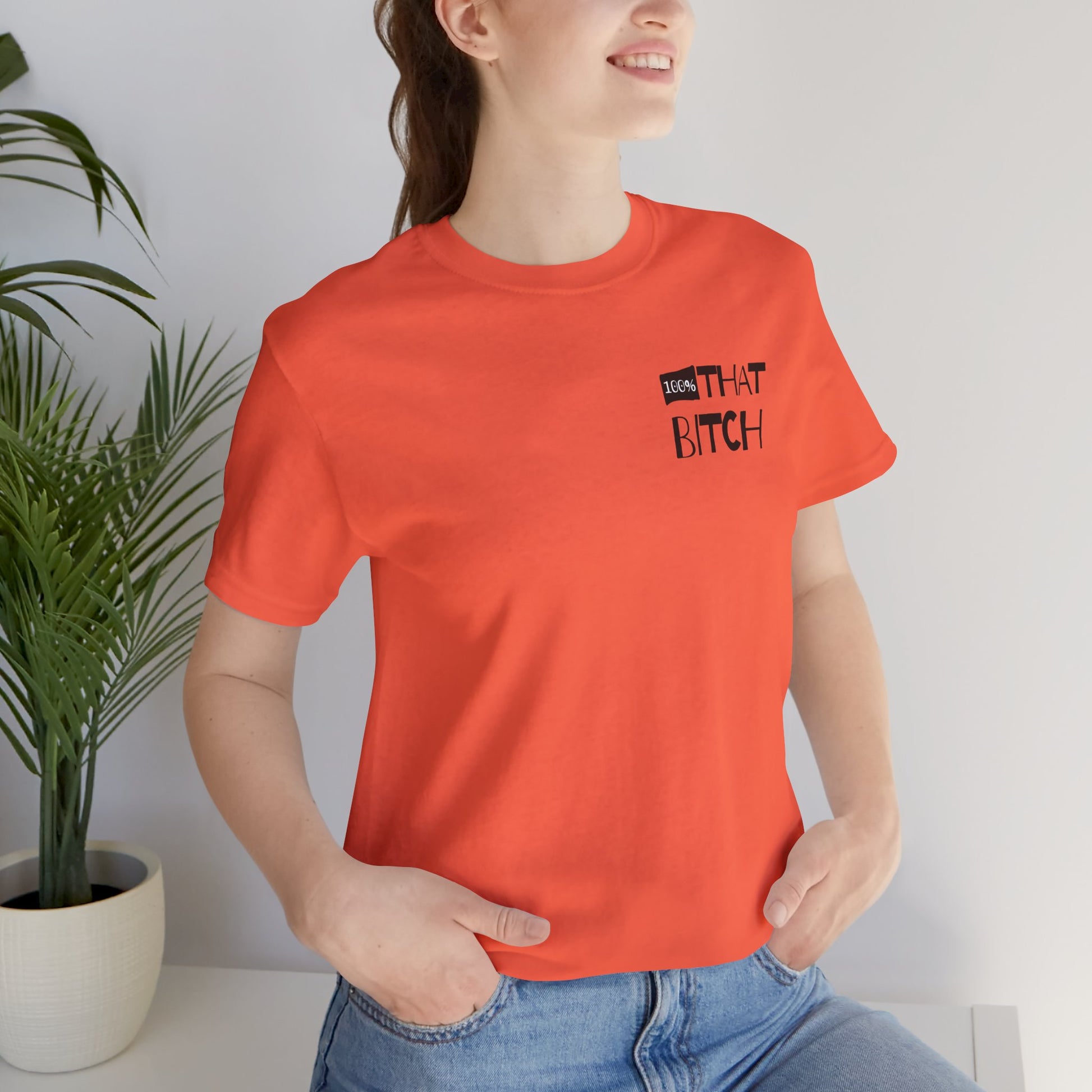 Person wearing an orange t-shirt with text, standing indoors next to a plant.  100% that Bitch