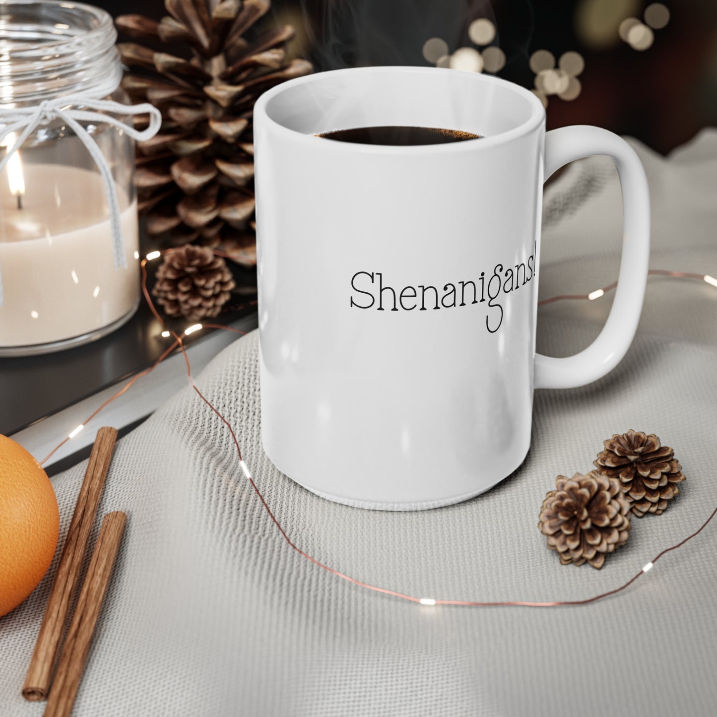 White mug with 'Shenanigans' text on a table with decorative items including a candle, oranges, and pine cones.