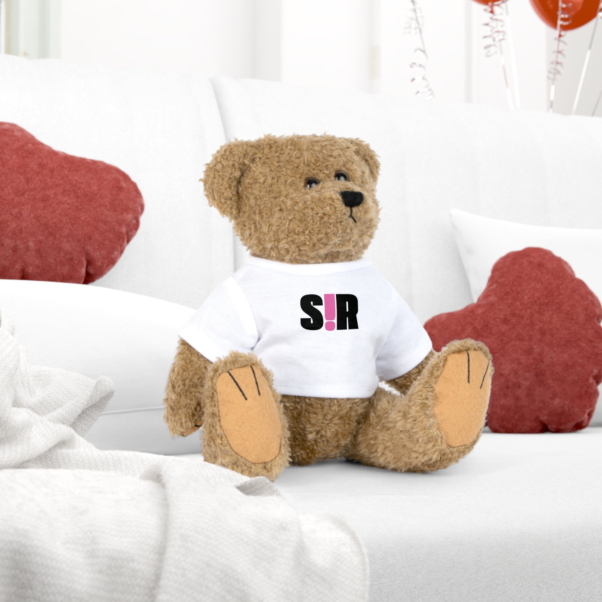 Brown teddy bear wearing a white shirt with 'SIR' printed on it, sitting on a white surface with red heart-shaped objects in the background.