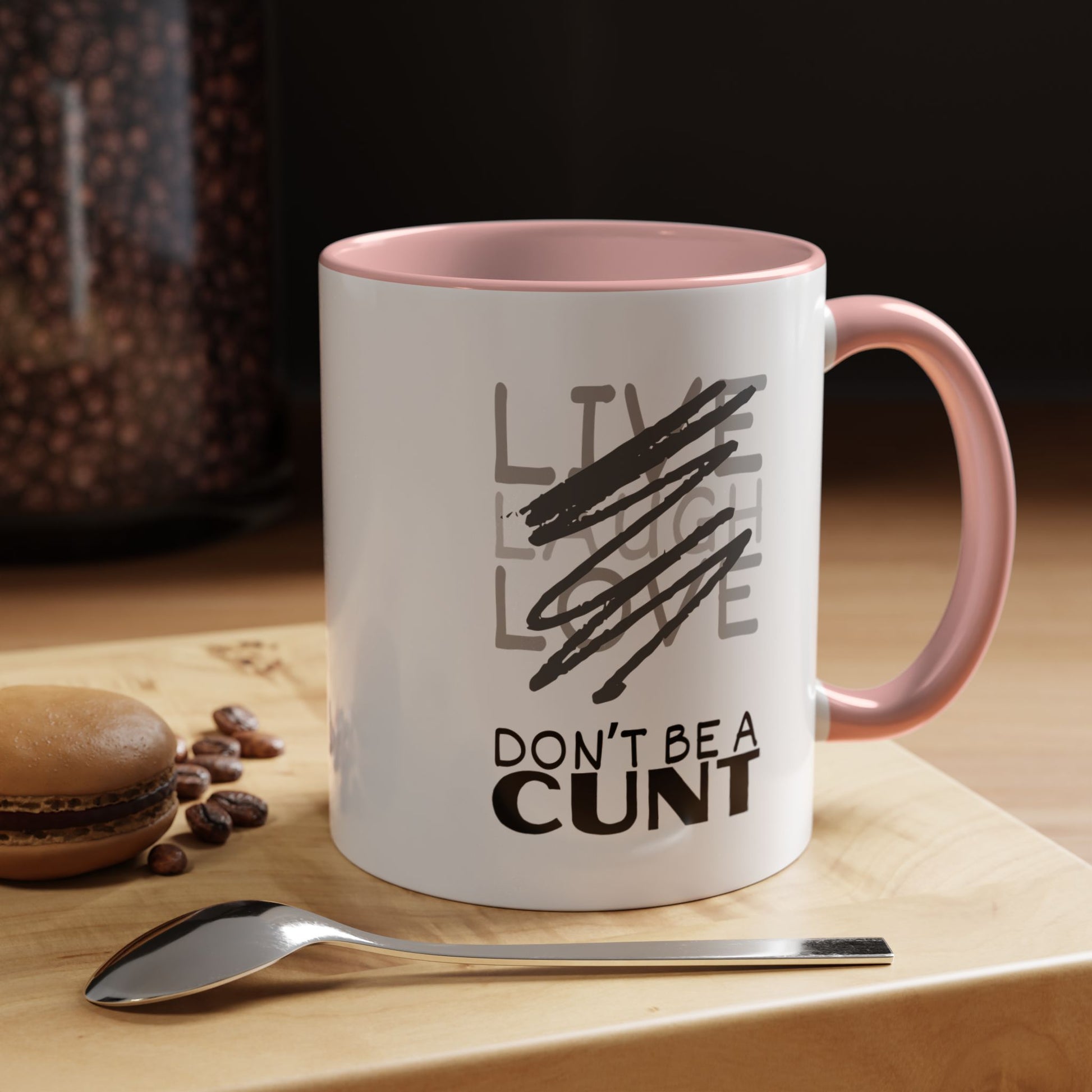 White mug with black text on a wooden surface with coffee beans and a spoon.