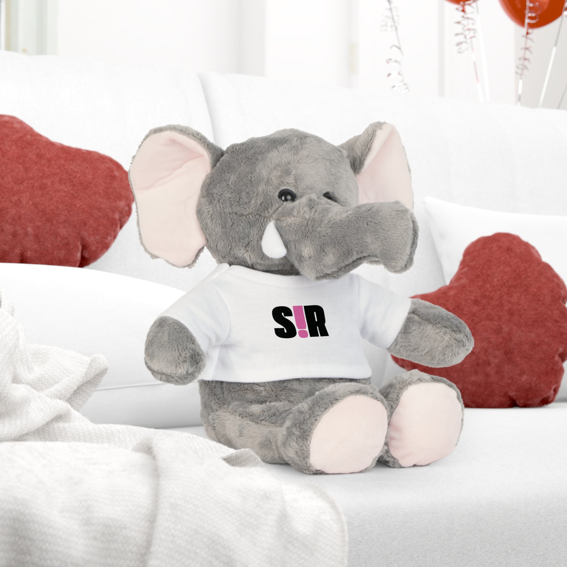 Gray stuffed elephant toy wearing a white shirt with 'SIR' printed on it, sitting on a bed with red heart-shaped pillows.