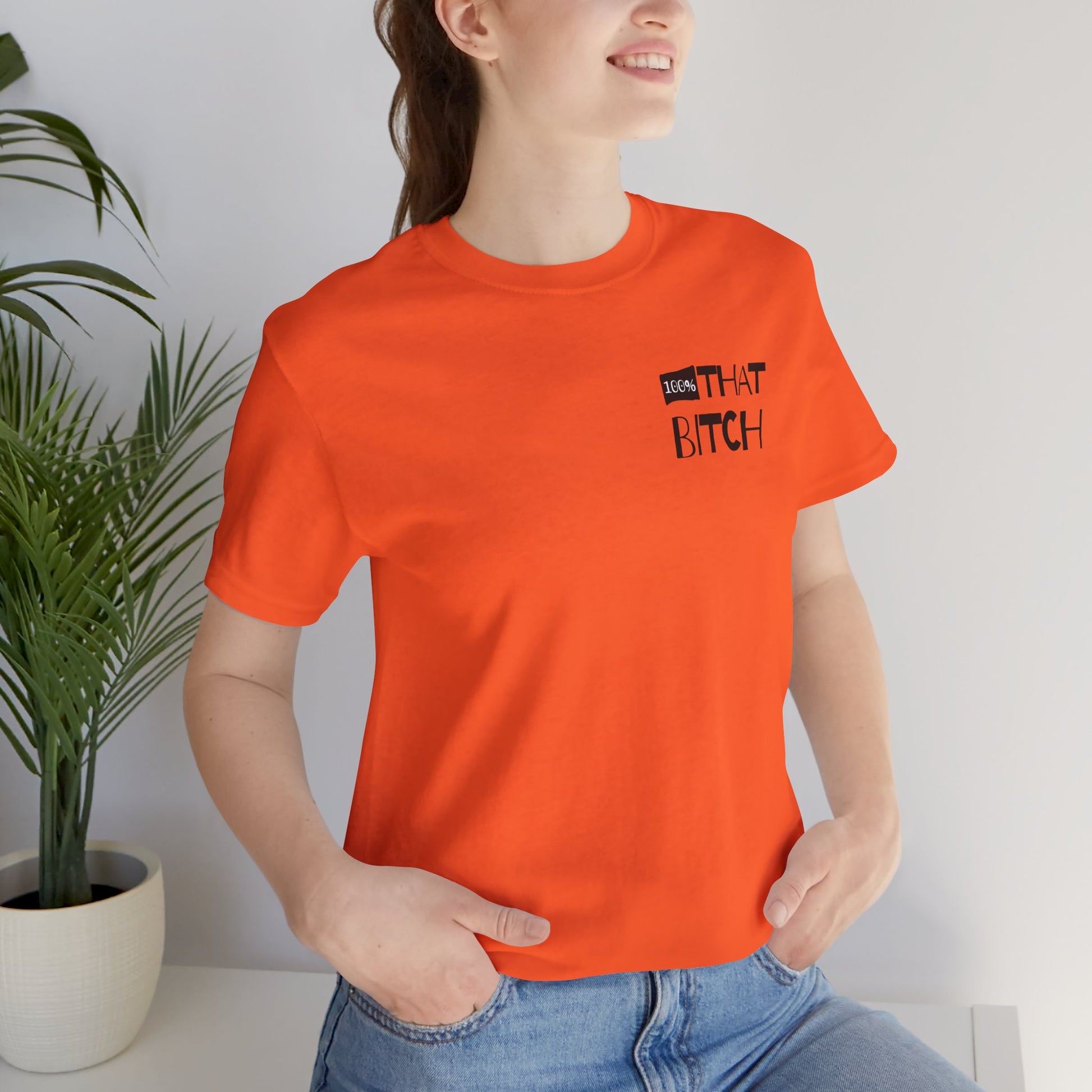 Person wearing an orange t-shirt with text, standing indoors next to a plant.  100% that Bitch