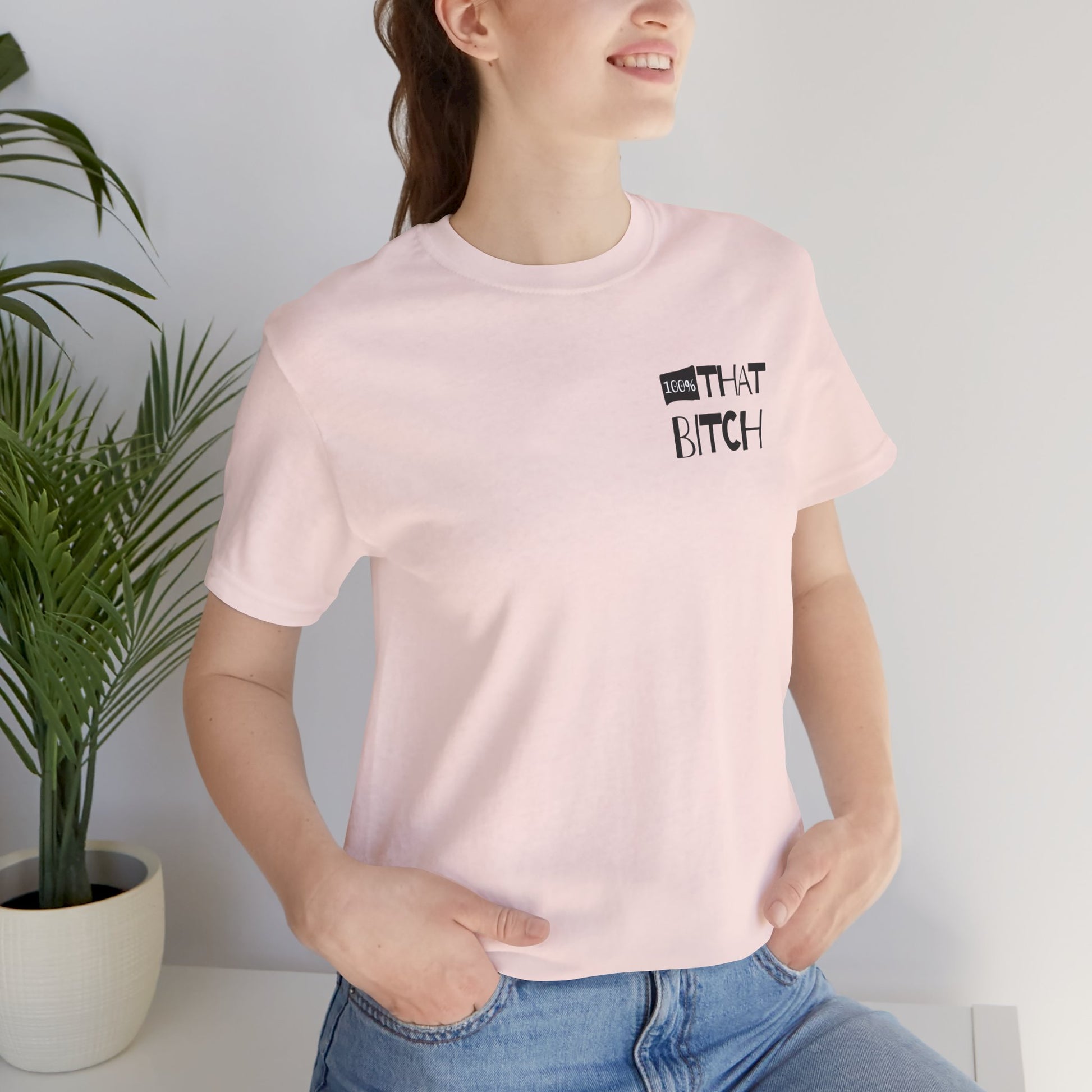 Person wearing a pink t-shirt with text, standing next to a plant indoors.  100% that Bitch