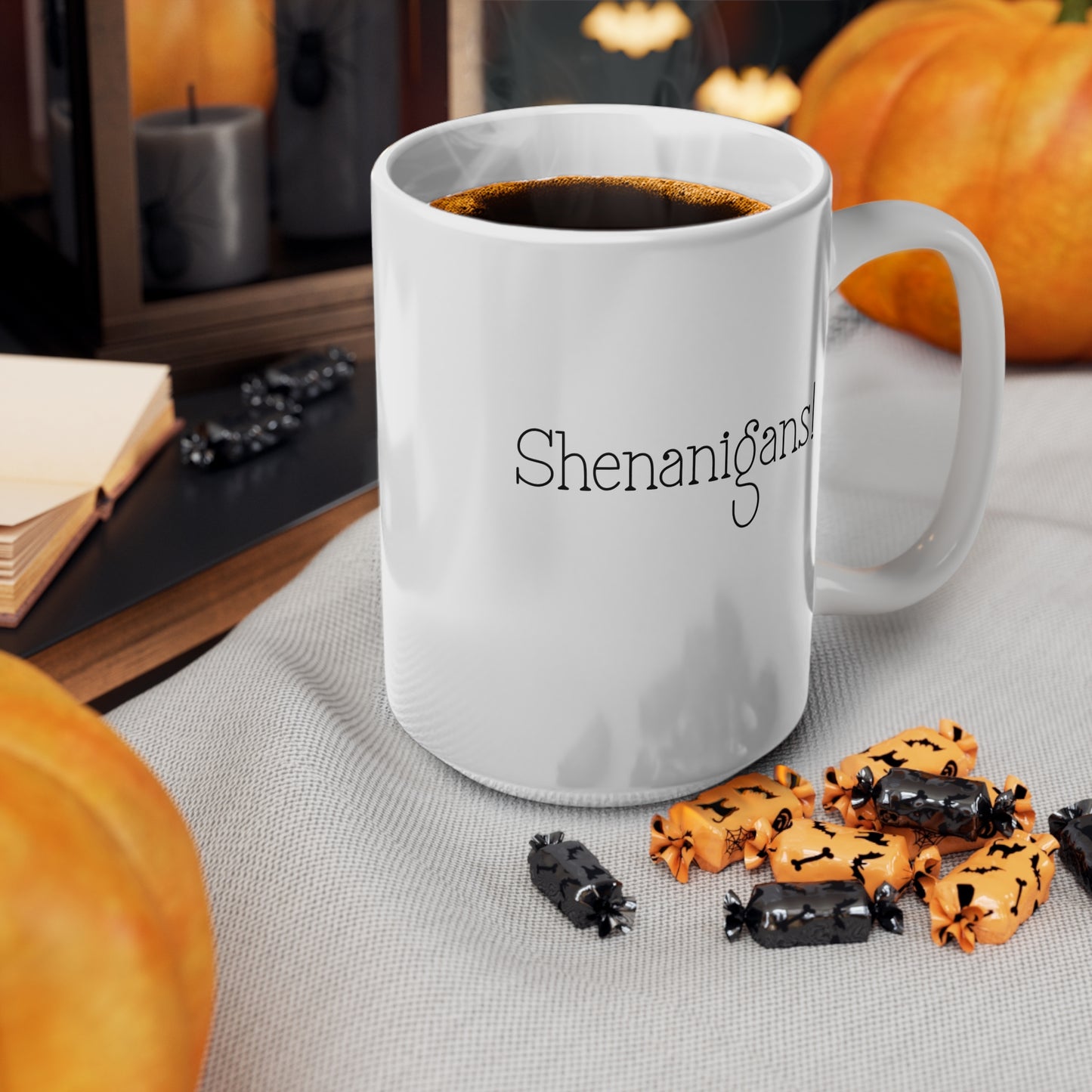 White mug with 'Shenanigans' text, filled with coffee, on a table with Halloween-themed cookies and pumpkins.
