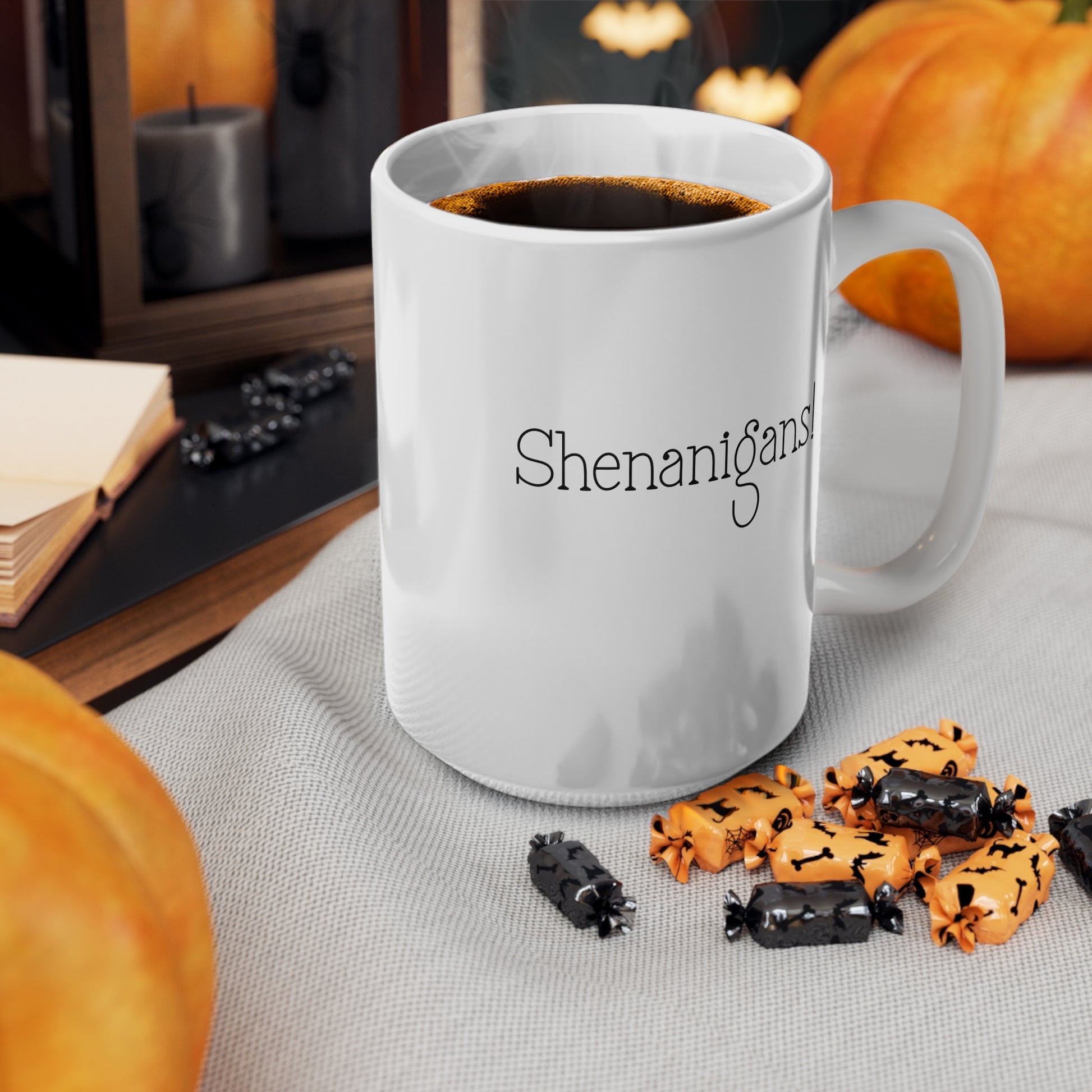 White mug with 'Shenanigans' text, filled with coffee, on a table with Halloween-themed cookies and pumpkins.