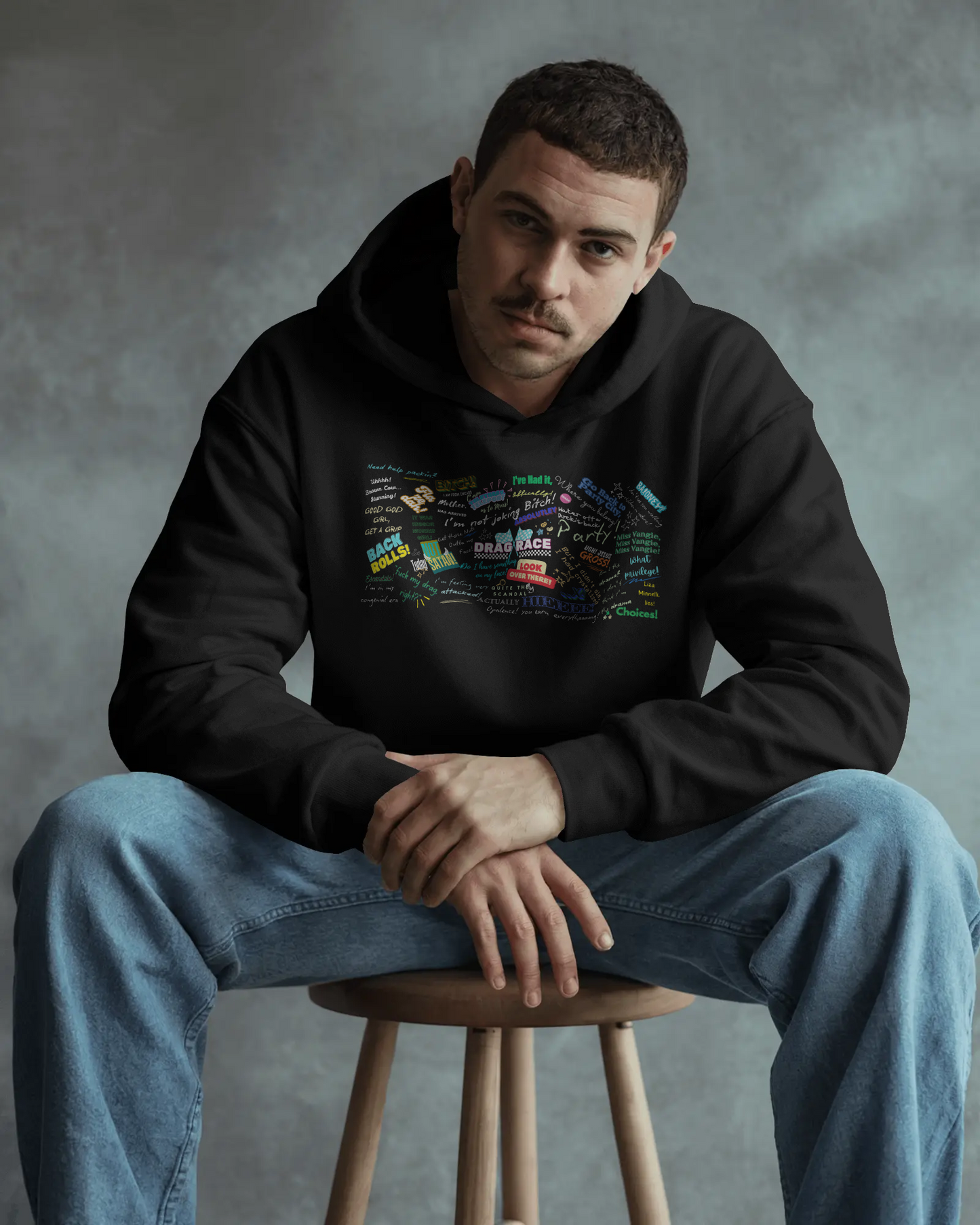 Man wearing a black hoodie with colorful graphics sitting on a stool against a gray background  Drag race, Rupaul, Rupaul's Drag Race UK, LGBT, Queer Culture, Gay, Drag Queen