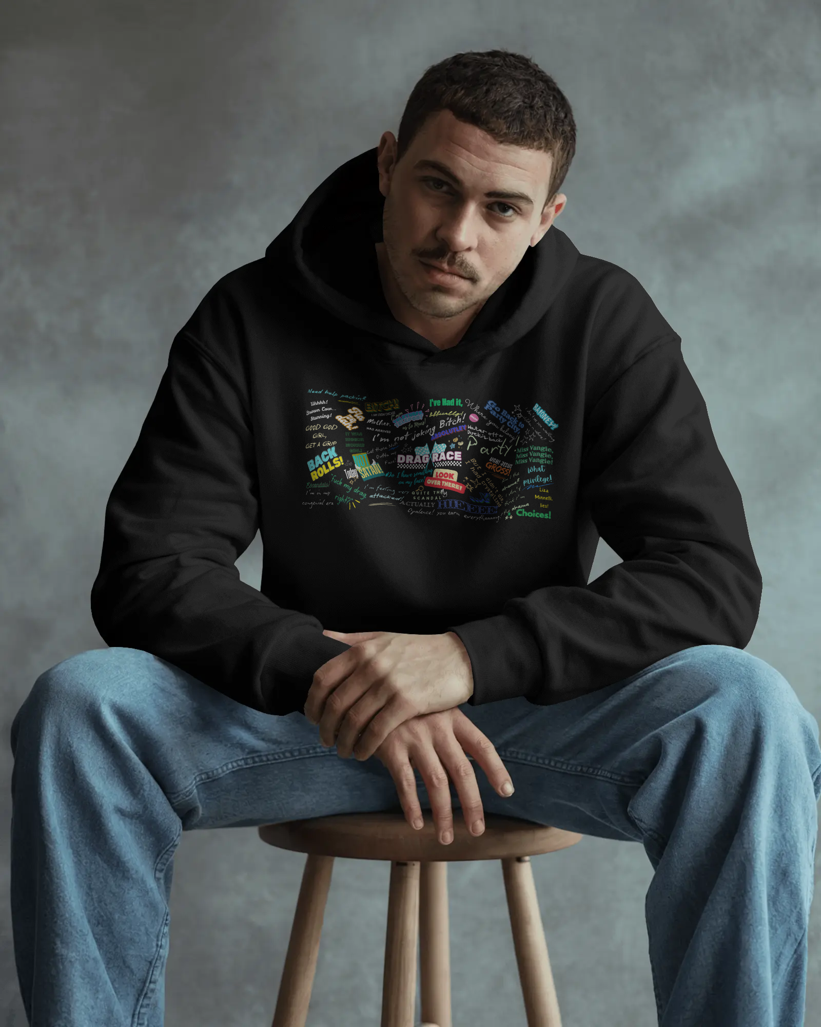 Man wearing a black hoodie with colorful graphics sitting on a stool against a gray background  Drag race, Rupaul, Rupaul's Drag Race UK, LGBT, Queer Culture, Gay, Drag Queen
