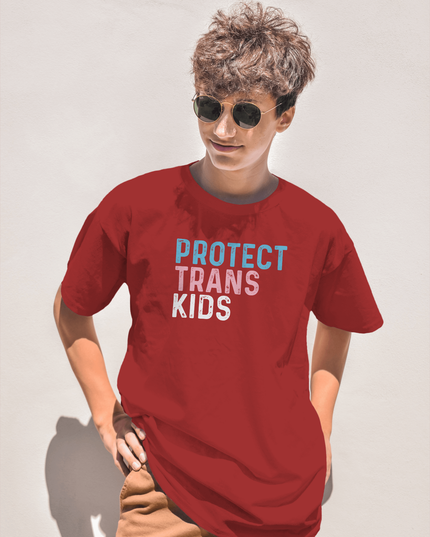 Person wearing a red t-shirt with 'PROTECT TRANS KIDS' text on a plain background LGBTQIA+, Queer, Trans rights