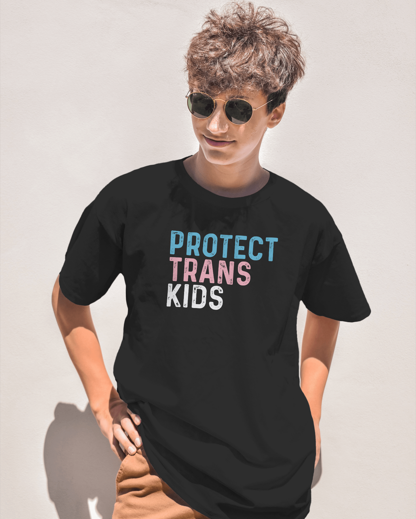 Person wearing a black t-shirt with 'PROTECT TRANS KIDS' text on a plain background LGBTQIA+, Queer, Trans rights