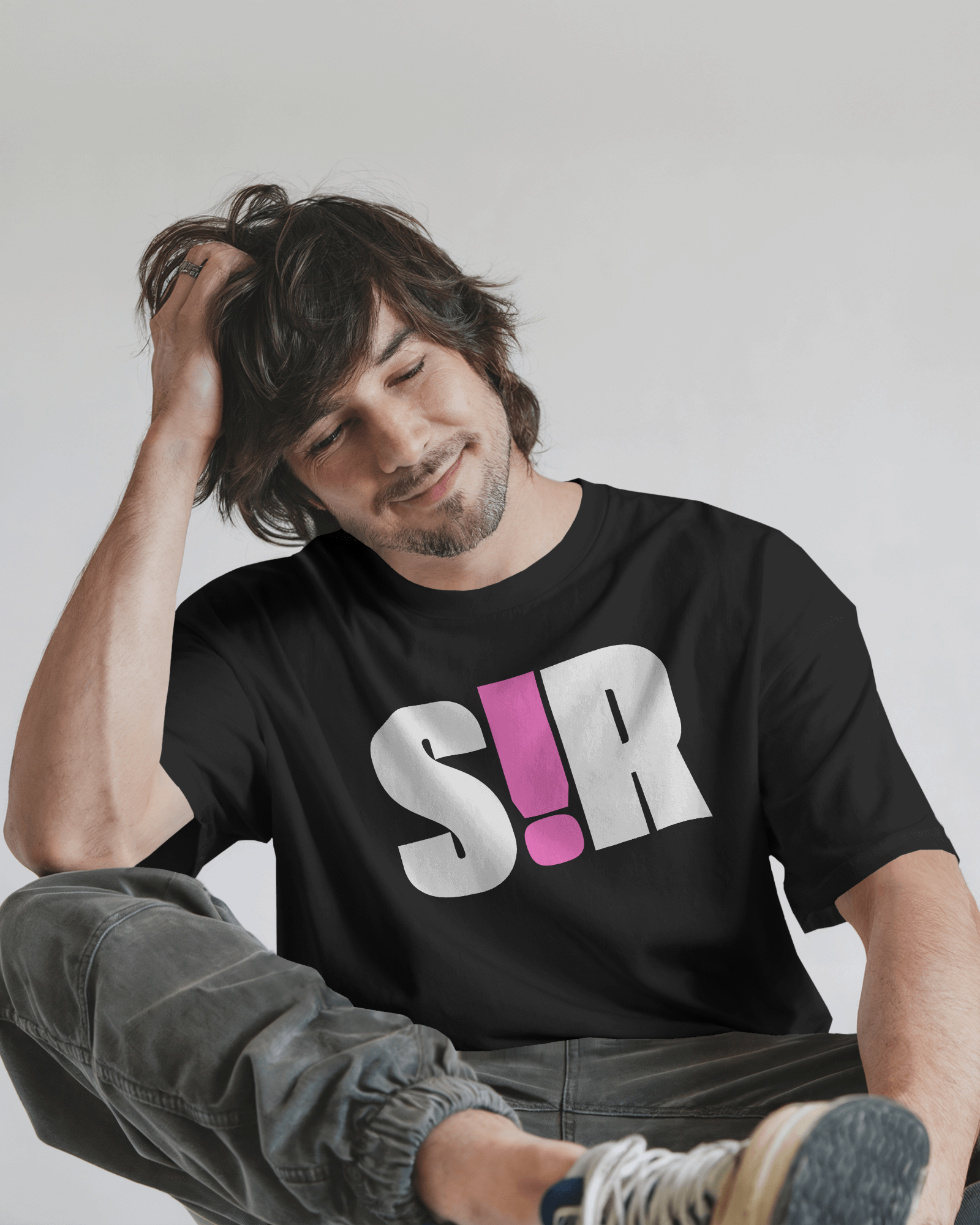 Man wearing a black t-shirt with 'S!R' printed on it, sitting against a plain background.  Kink, LGBTQIA+, Queer Sub Dom