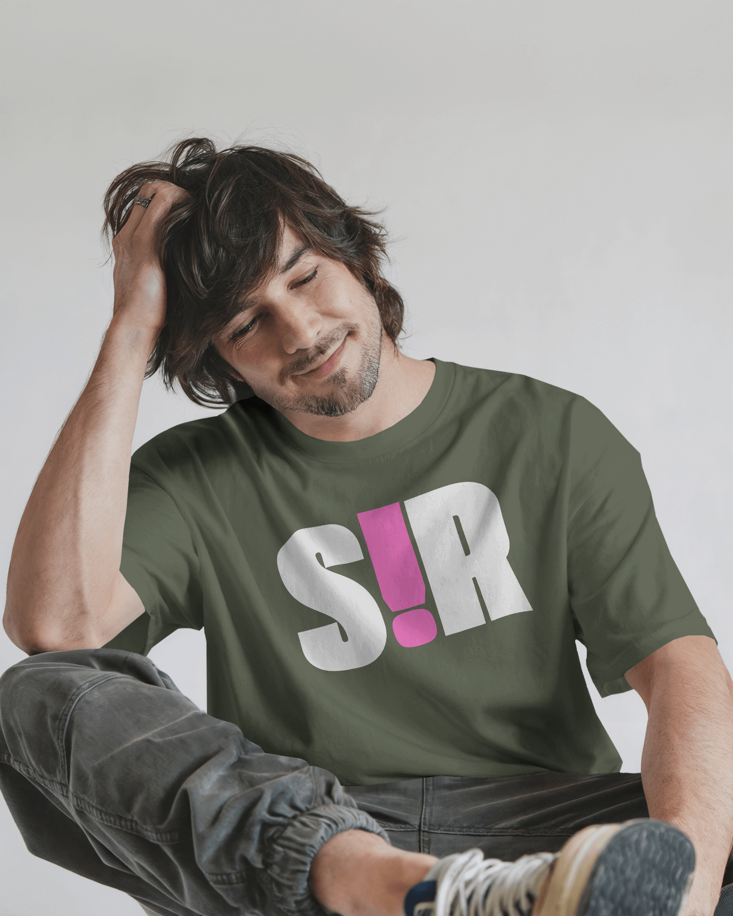 Man wearing a green t-shirt with 'S!R' printed on it, sitting against a plain background. Kink, LGBTQIA+, Queer Sub Dom