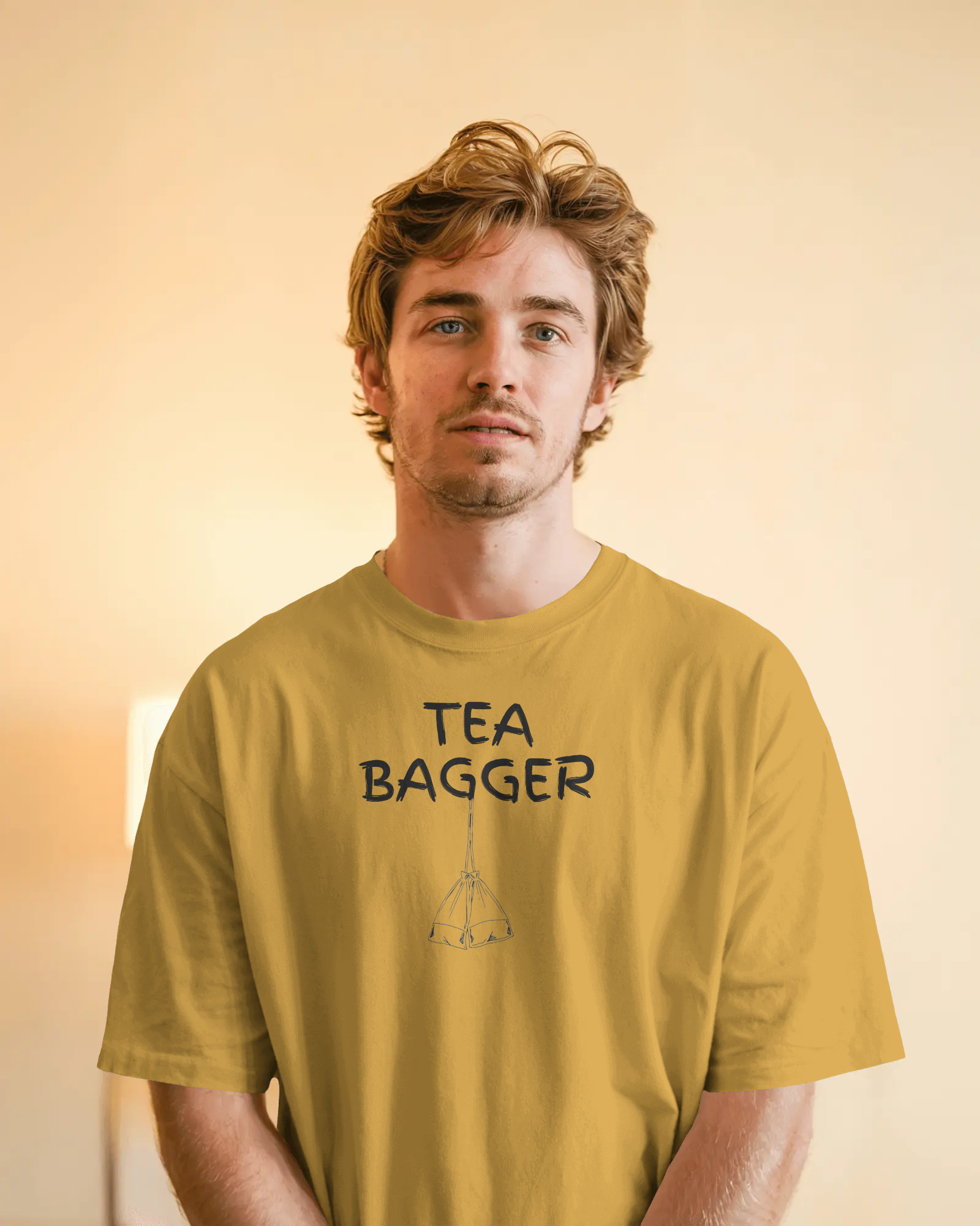 Man wearing a mustard yellow t-shirt with 'TEA BAGGER' text and graphic on a plain background  Kink, LGBTQIA+, Queer