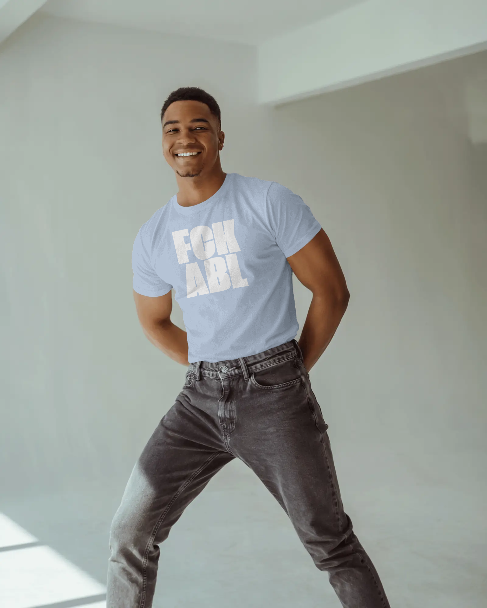 Man wearing a light blue t-shirt with text and dark jeans standing in a well-lit room.