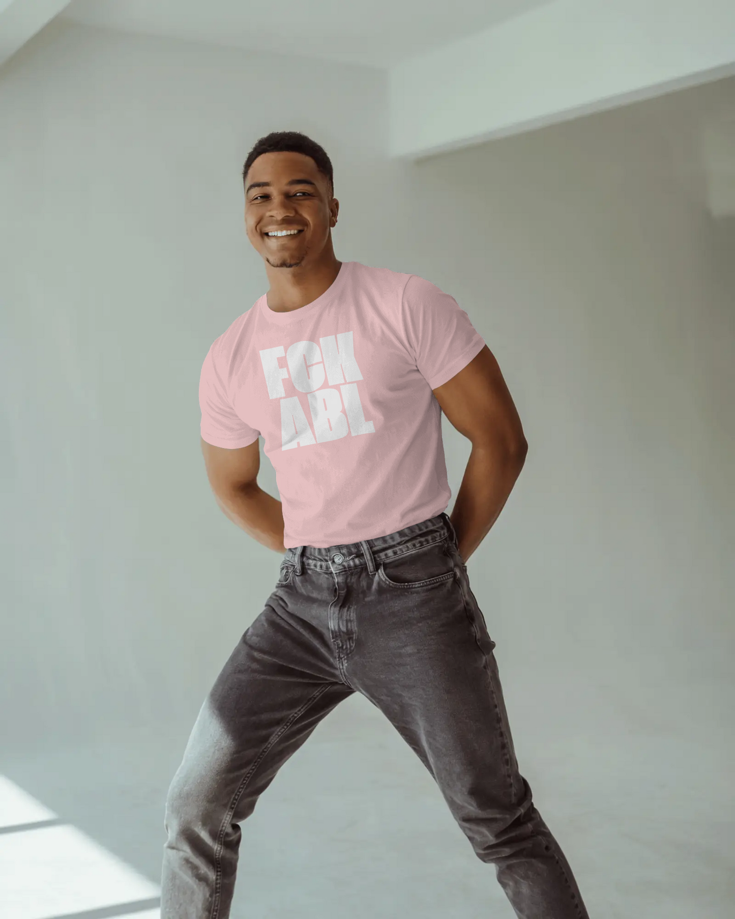 Man wearing a pink t-shirt with text and gray jeans standing against a white wall.