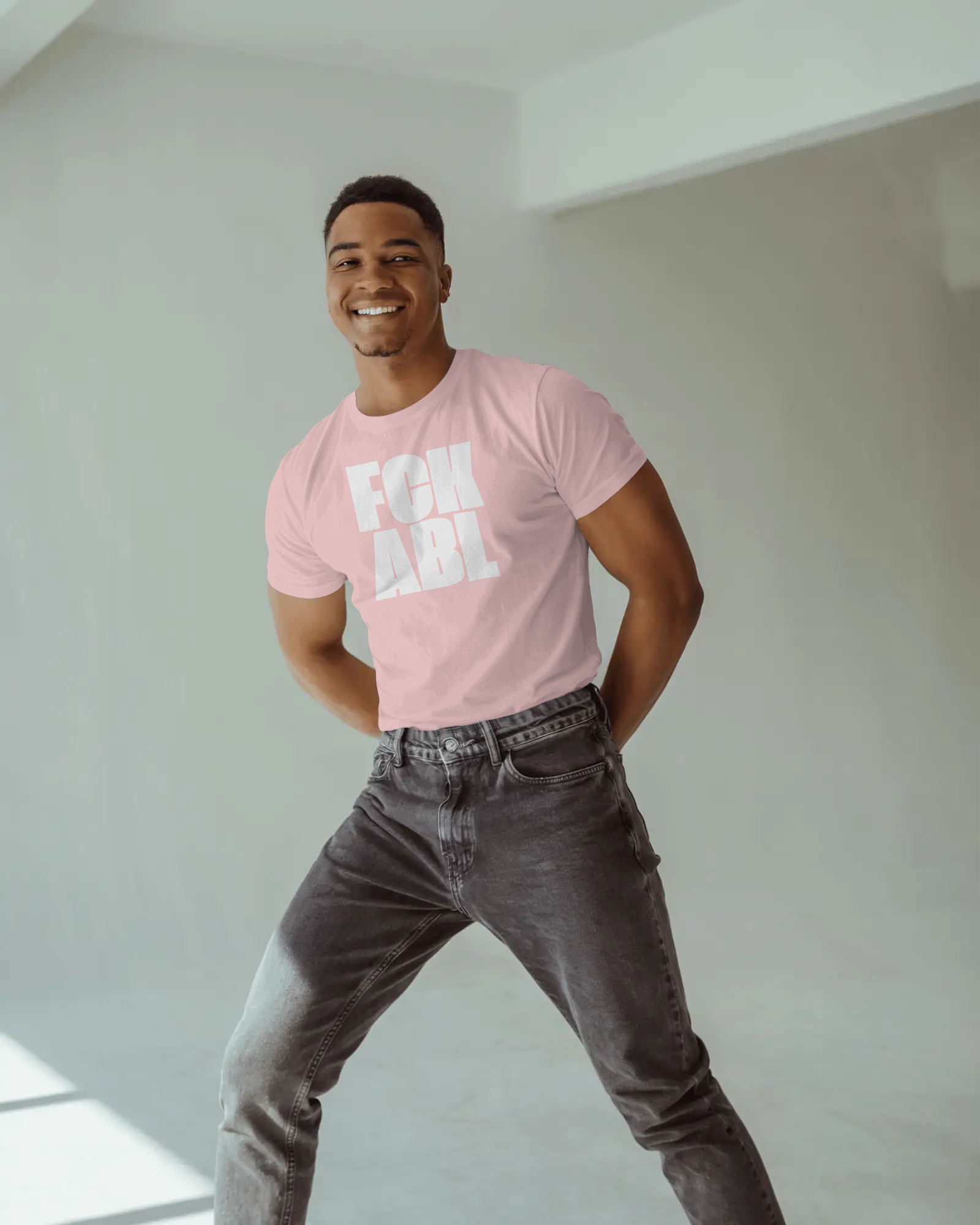 Man wearing a pink t-shirt with text and gray jeans standing against a white wall.