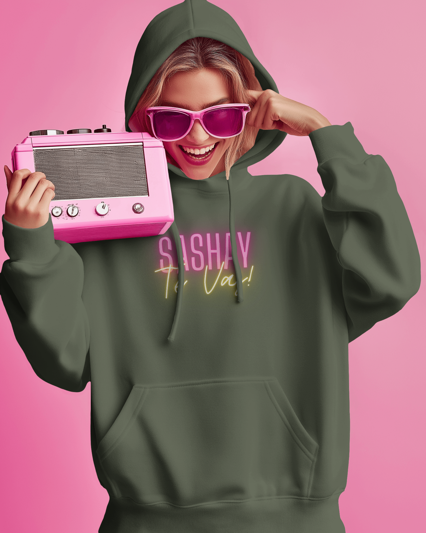 Person wearing a green hoodie with text, holding a pink vintage radio against a pink background  Drag Race Espana, Drag Race Spain, Rupaul's drag race, Drag queens, LGBT, Queer culture