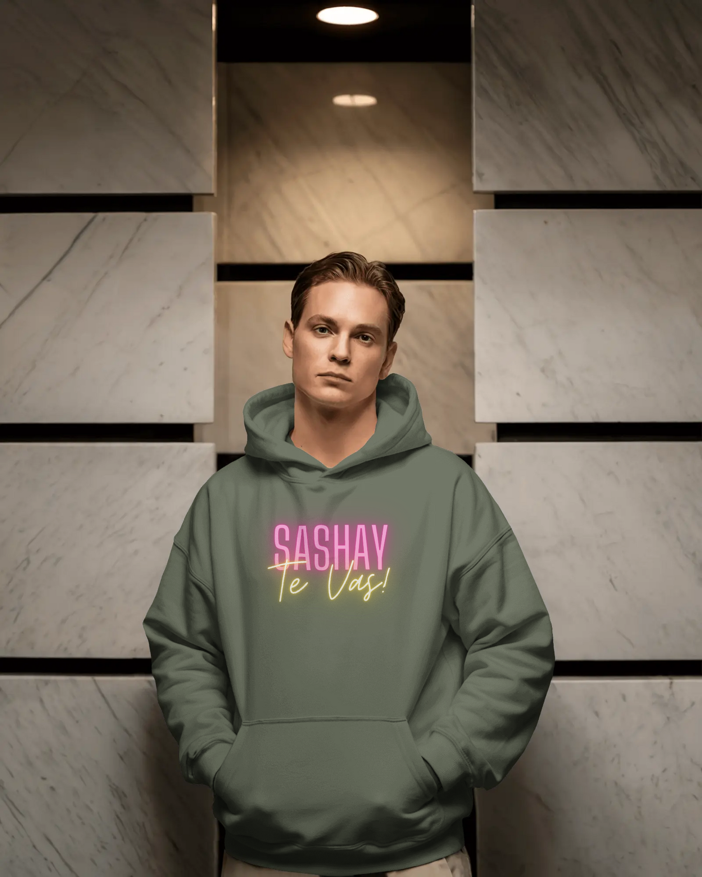 Person wearing a green hoodie with 'SASHAY Te Vas! text in pink and yellow, standing against a neutral wall.