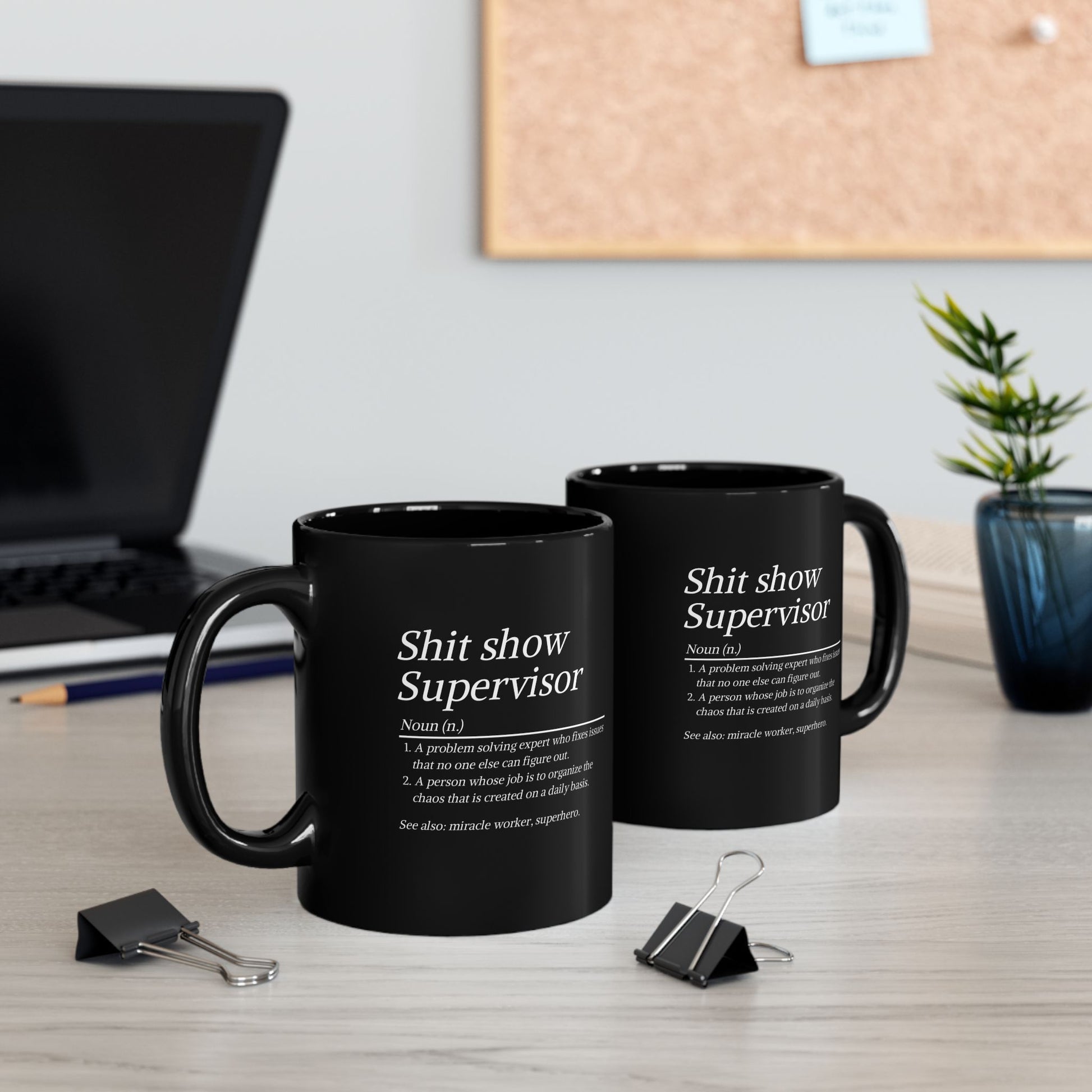 Two black mugs with humorous text on a desk with office supplies and decor.