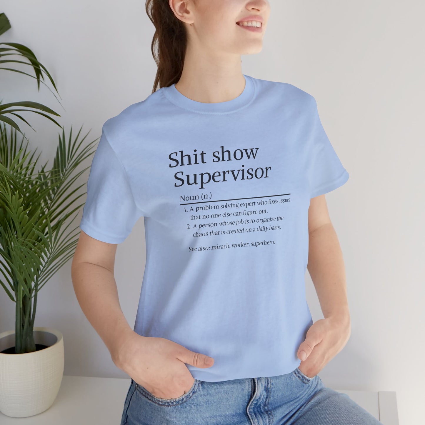 Person wearing a light blue t-shirt with text, standing indoors next to a plant.