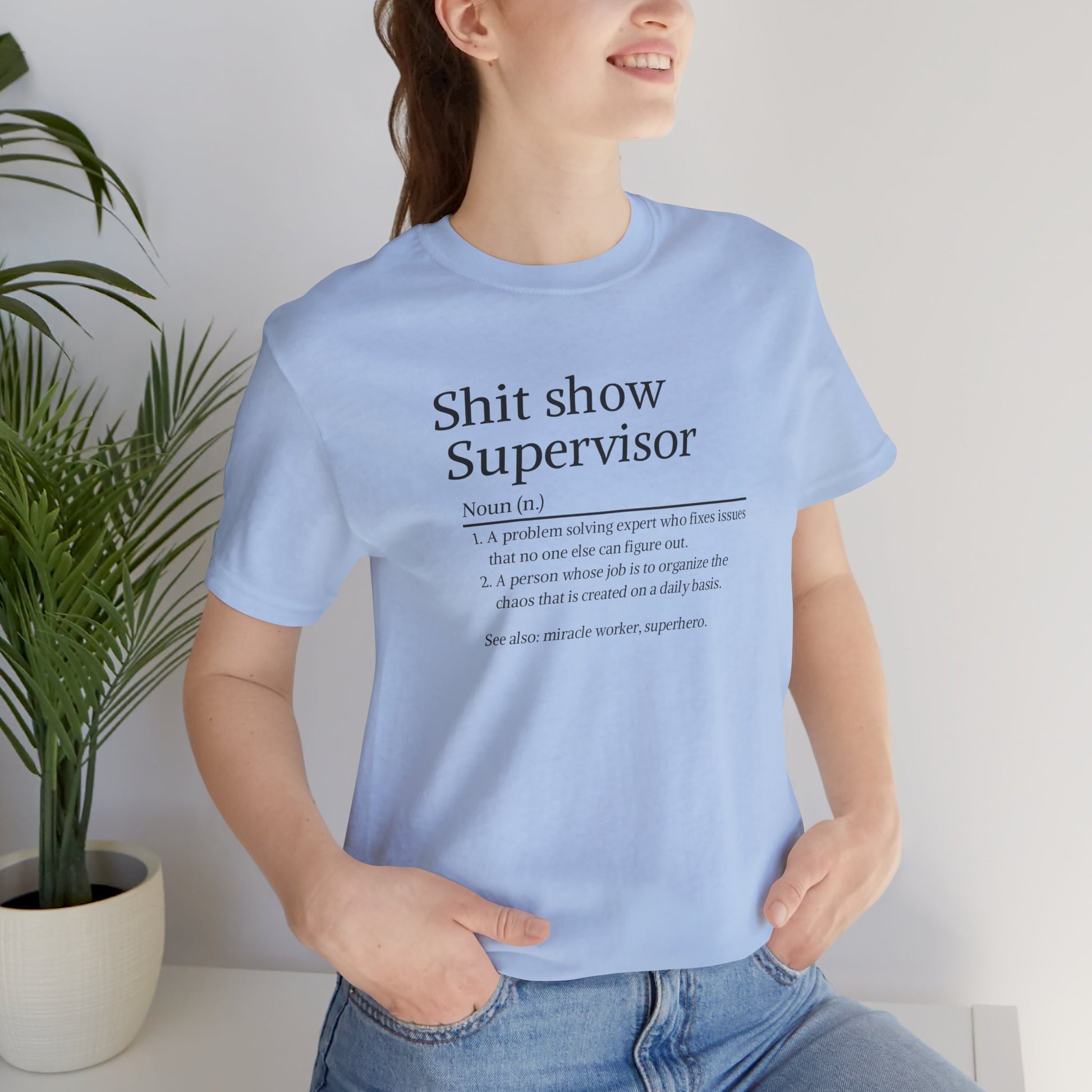 Person wearing a light blue t-shirt with text, standing indoors next to a plant.