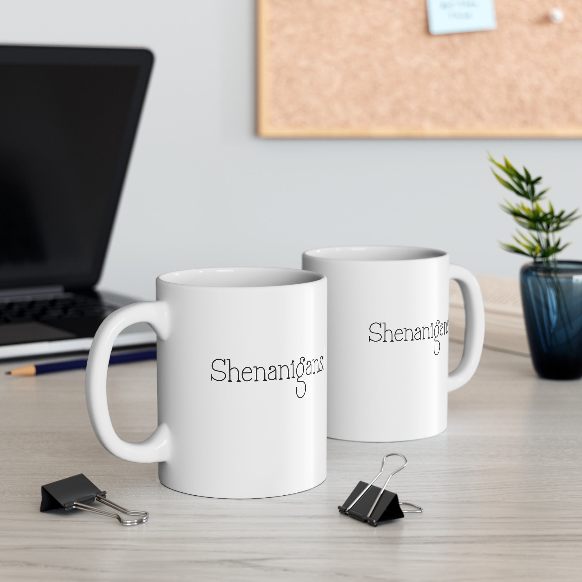 Two white mugs with 'Shenanigans' text on a desk with office items.