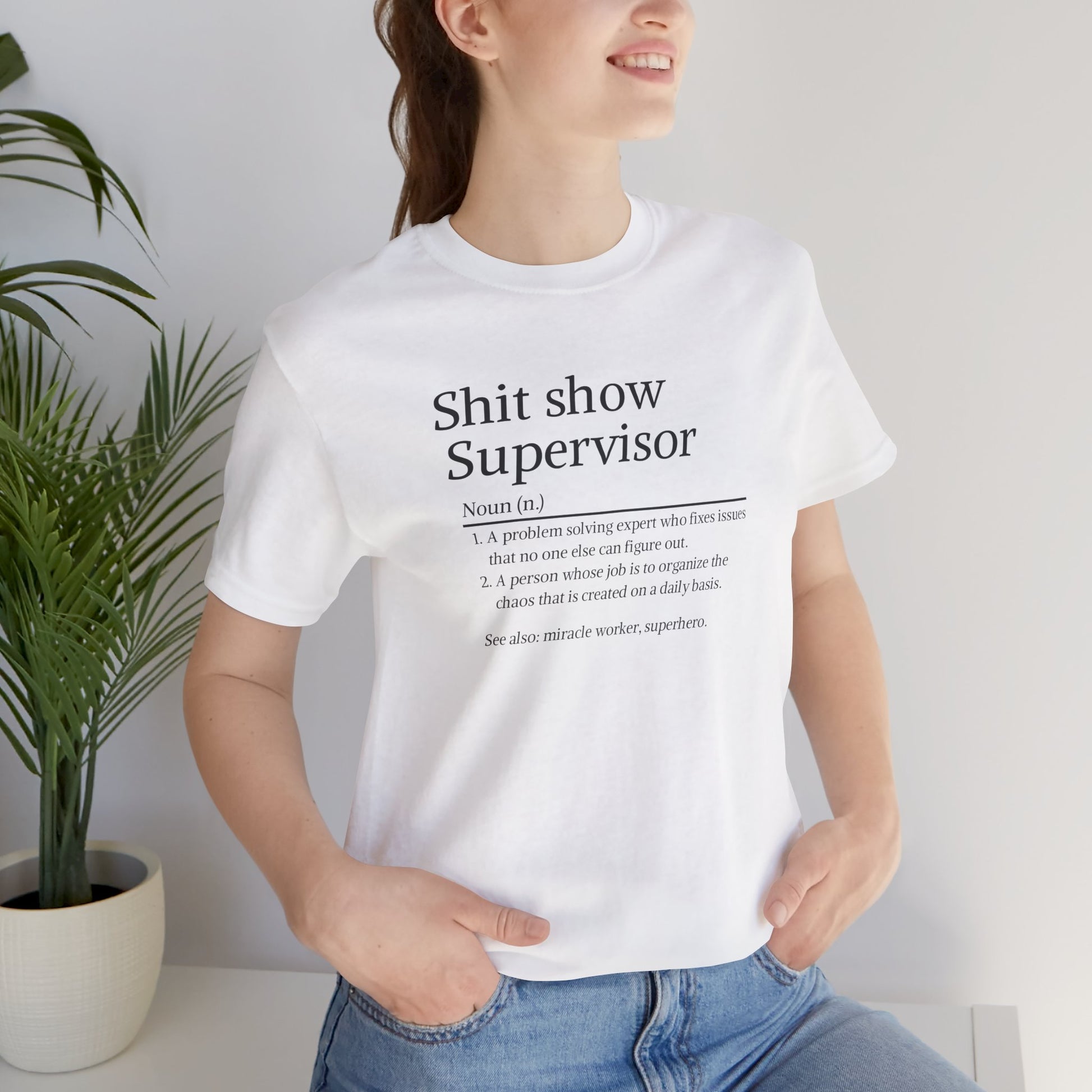 Person wearing a white t-shirt with text, standing indoors next to a plant.